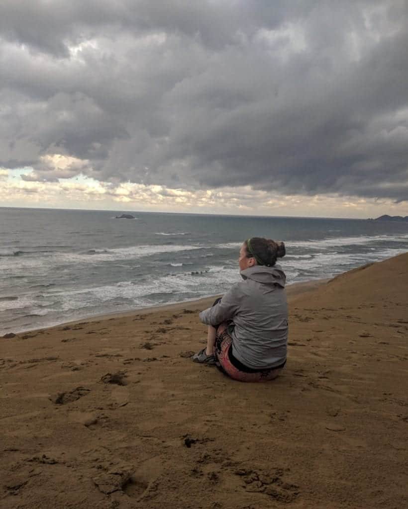 A young woman sits with a wind jacket on top of the 100 feet tall Tottori Sand Dunes with a stormy sky in the background