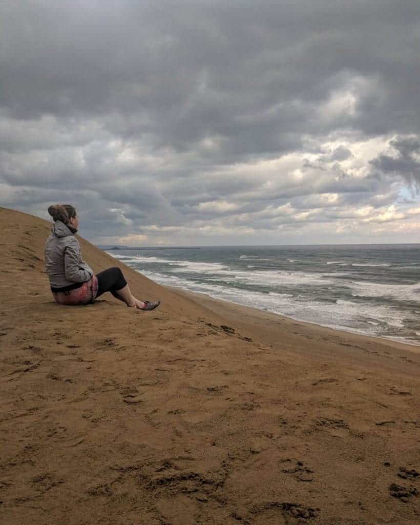 A young woman sits with a wind jacket on top of the 100 feet tall Tottori Sand Dunes with a stormy sky in the background